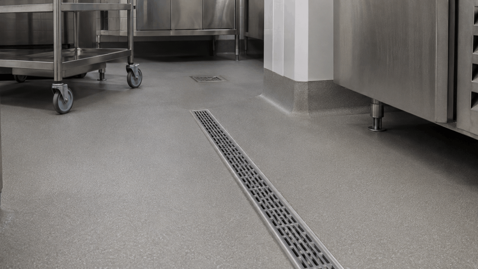 Seamless grey epoxy commercial kitchen floor with a stainless steel trolley, linear floor drain, and equipment